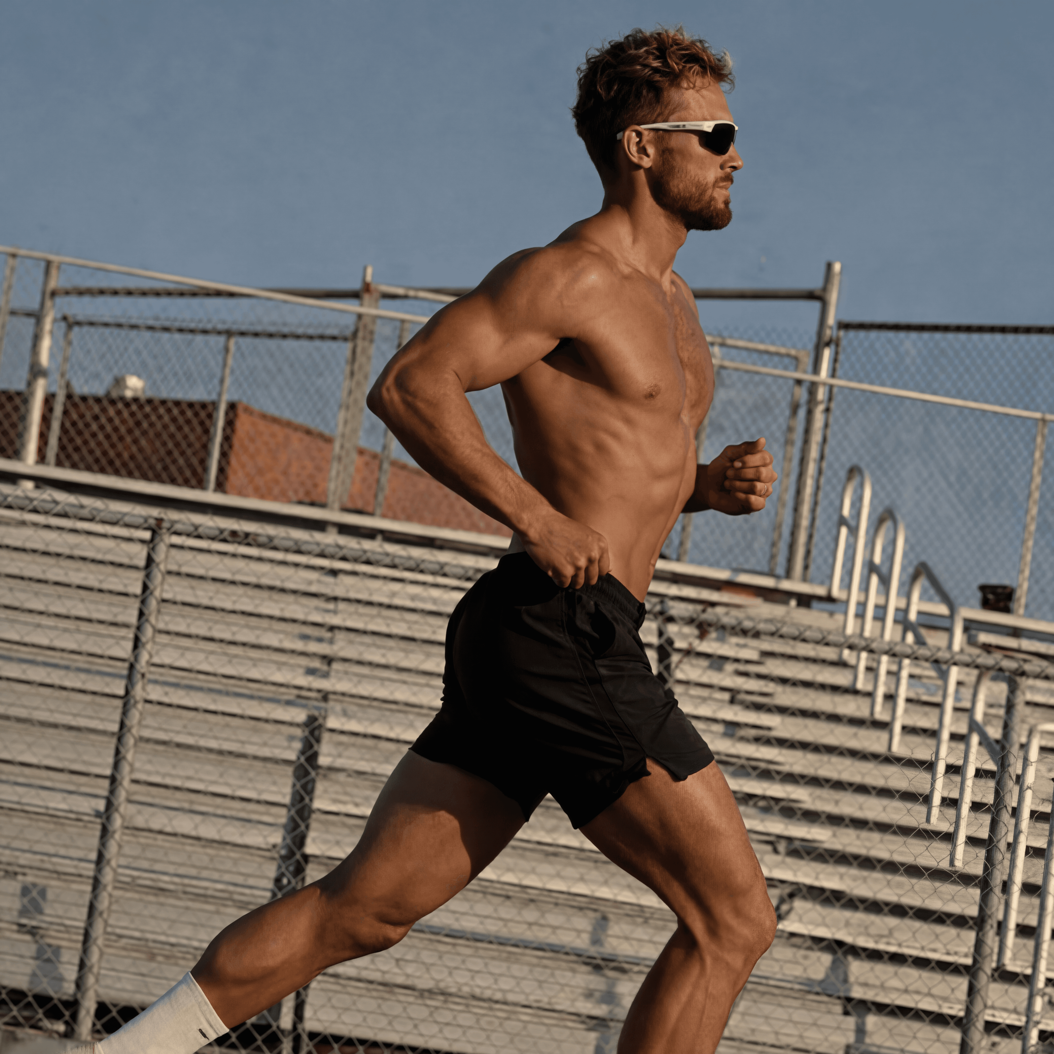 Man running shirtless outdoors on a track, wearing black shorts and sunglasses, mid-stride in a focused workout.