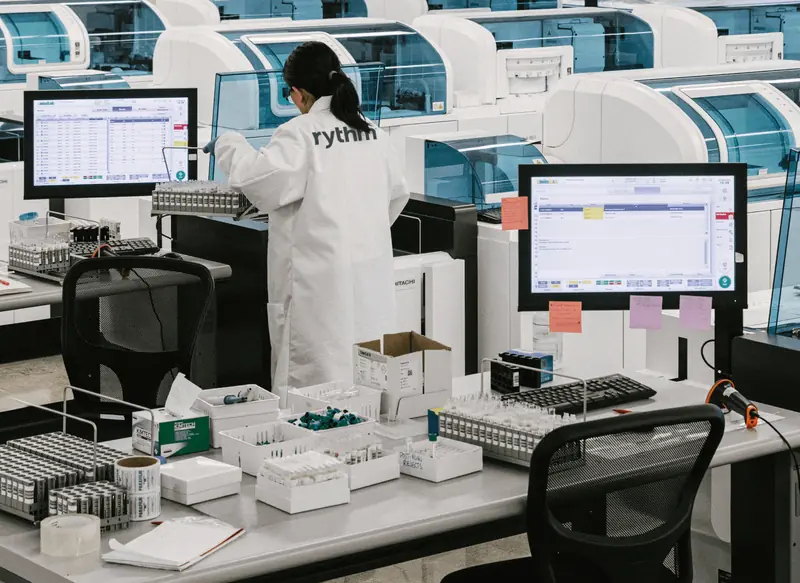 Laboratory technician working with automated testing machines and sample trays in a clinical lab environment.