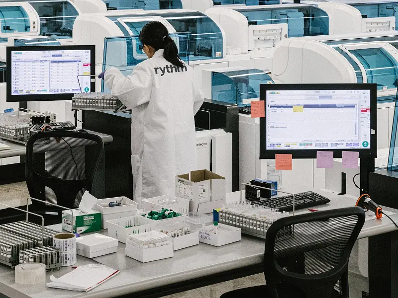 Lab technician in a Rythm lab processing blood samples at a workstation, surrounded by testing equipment, vials, and diagnostic machines.