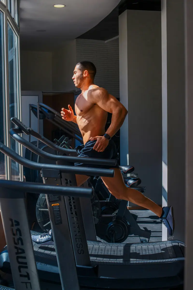 Man running on a treadmill in a modern gym with sunlight coming through large windows.