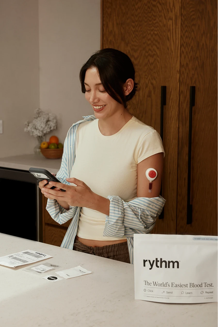 Woman using a Rythm at-home blood collection device on her upper arm, seated indoors in soft natural light, looking down as she applies the device.