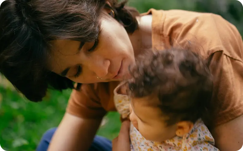 Mother tenderly leaning over her young child outdoors in a lush green setting.