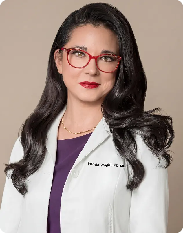 Female doctor wearing a white coat and red glasses, with long dark hair and a composed expression, photographed against a clean studio background.