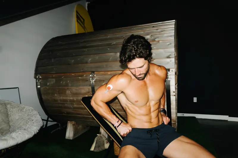 Shirtless athletic man sitting near a sauna, looking down at a Rythm Health sensor on his upper arm.