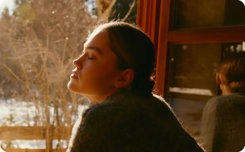 Woman with eyes closed basking in warm sunlight by a window, surrounded by a snowy winter landscape.