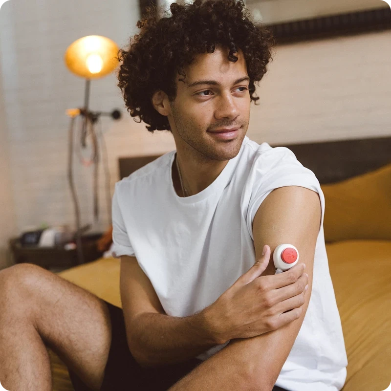 Smiling man sitting on a couch at home with a Rythm blood collection device adhered to his upper arm, in a bright, plant-filled living space.