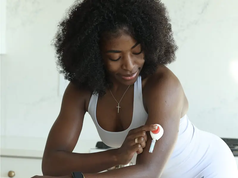 Woman with curly hair in athletic wear adjusting a Rythm Health sensor on her upper arm in a bright indoor space.
