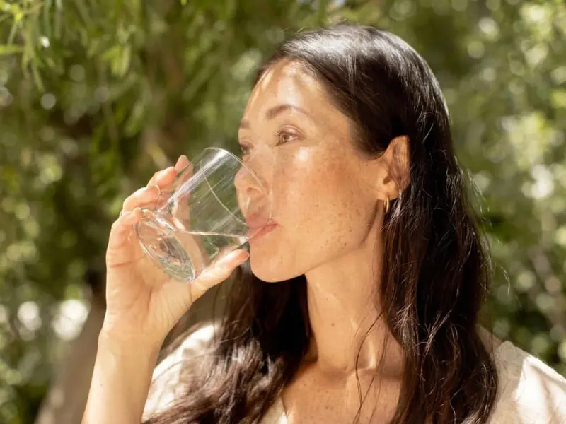 Woman drinking water outdoors in sunlight, emphasizing hydration and wellness.