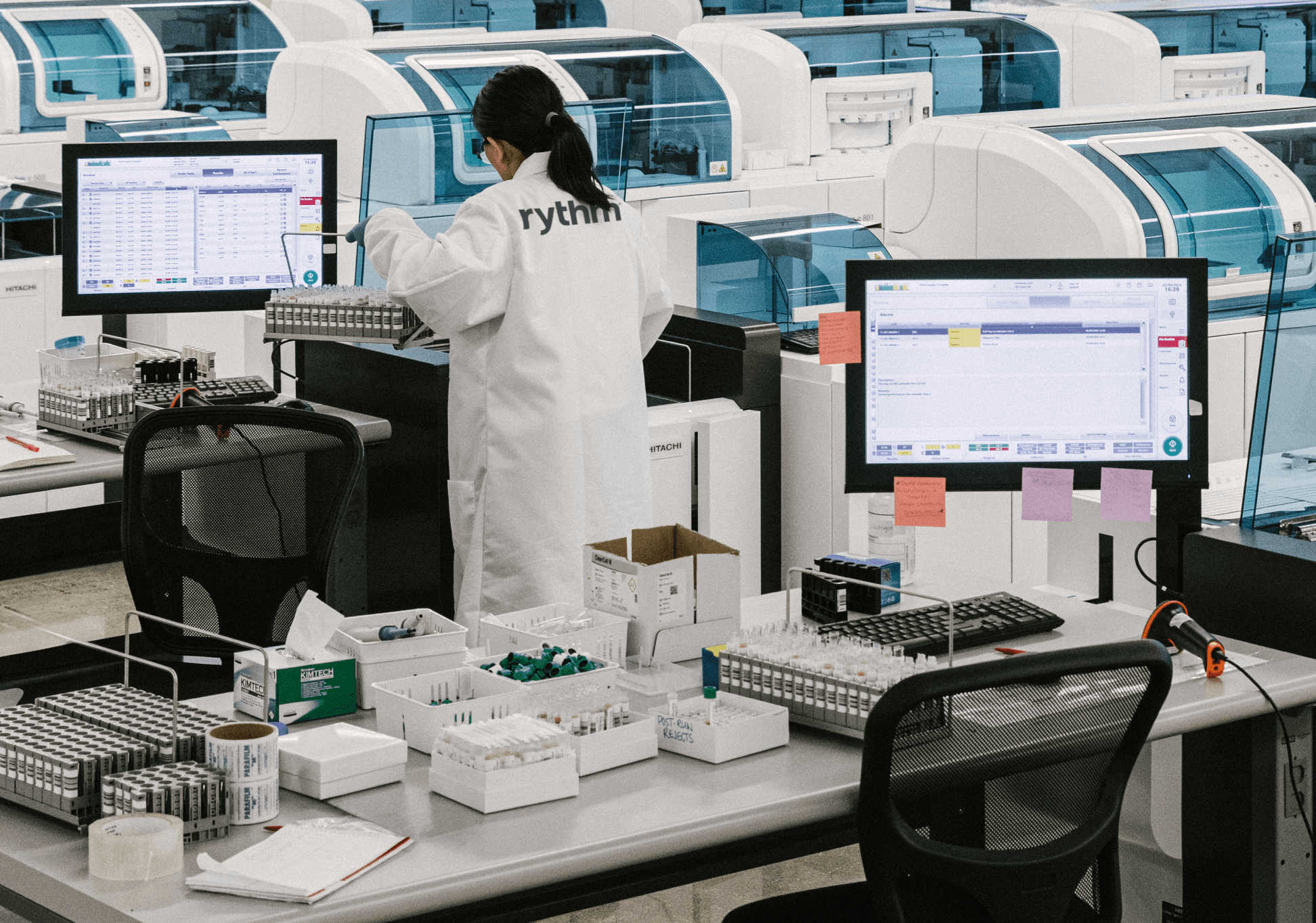 Laboratory technician working with automated testing machines and sample trays in a clinical lab environment.