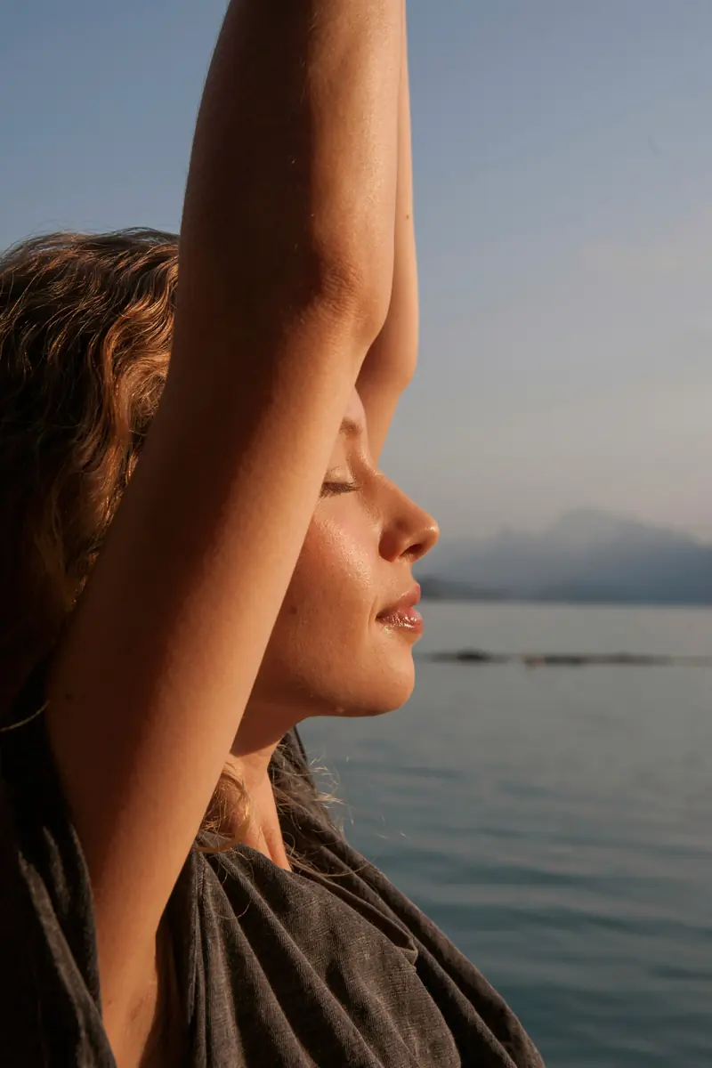 Woman stretching with eyes closed near the ocean at sunset, representing relaxation and recovery.