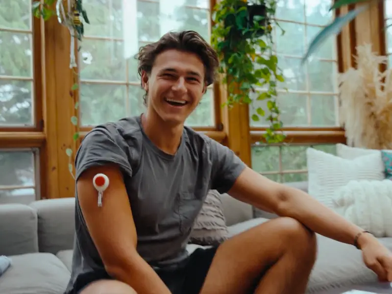 Smiling man sitting on a couch at home with a Rythm blood collection device adhered to his upper arm, in a bright, plant-filled living space.