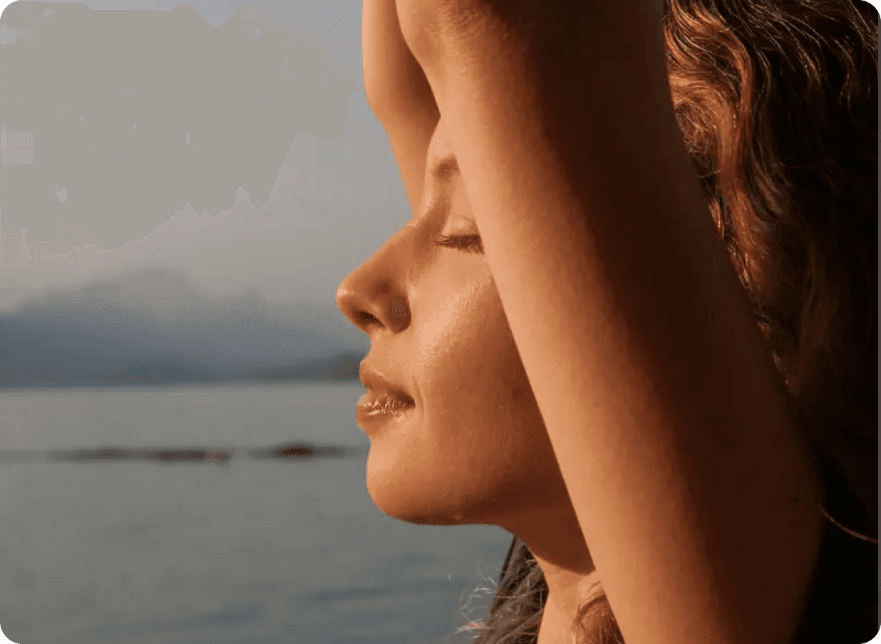 Woman stretching with eyes closed near the ocean at sunset, representing relaxation and recovery.