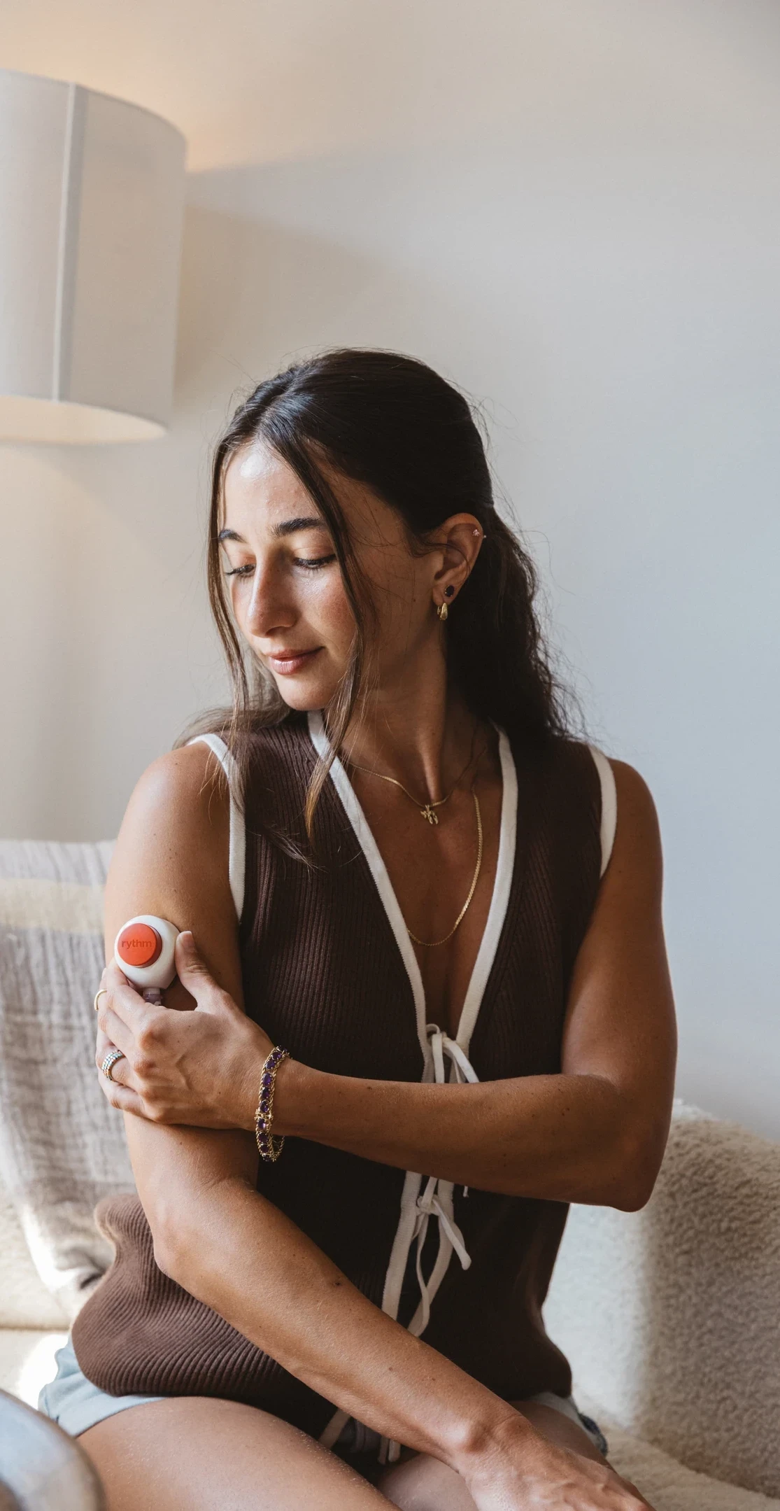 Woman using a Rythm at-home blood collection device on her upper arm, seated indoors in soft natural light, looking down as she applies the device.