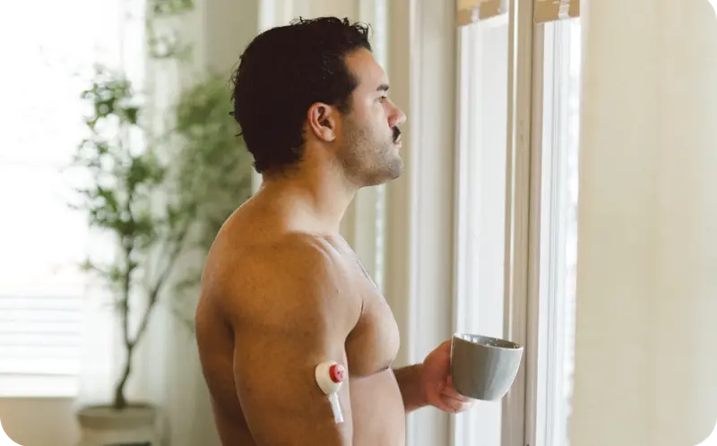 Shirtless man holding a coffee mug and gazing out a window with a Rythm Health device applied to his arm.