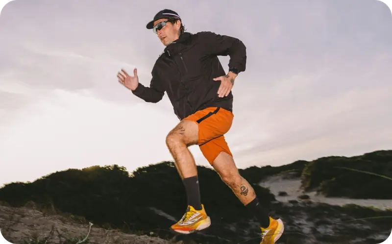 Male trail runner in a black jacket and orange shorts sprinting across a coastal dune at dusk.