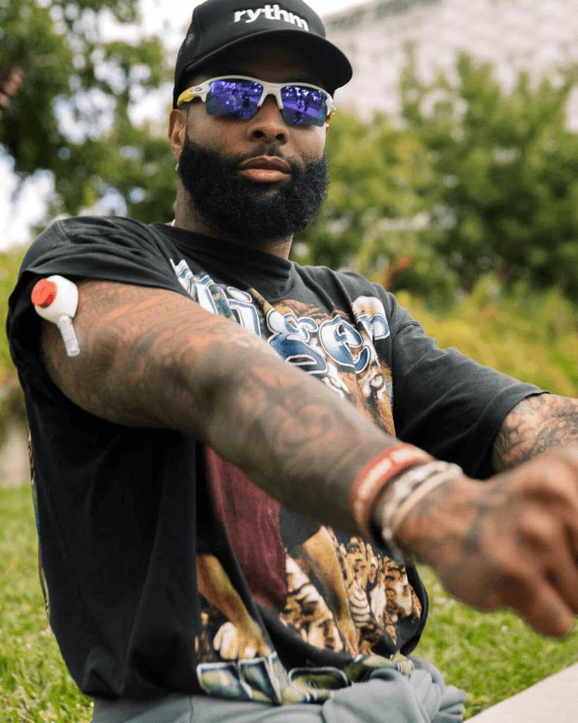 Man wearing sunglasses and a black cap outdoors, showing a Rythm device applied to his upper arm.